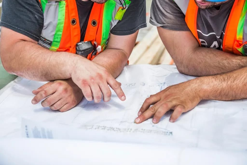 Two site workers in high-visibility vests leaning over a printed drawing and pointing at a detail