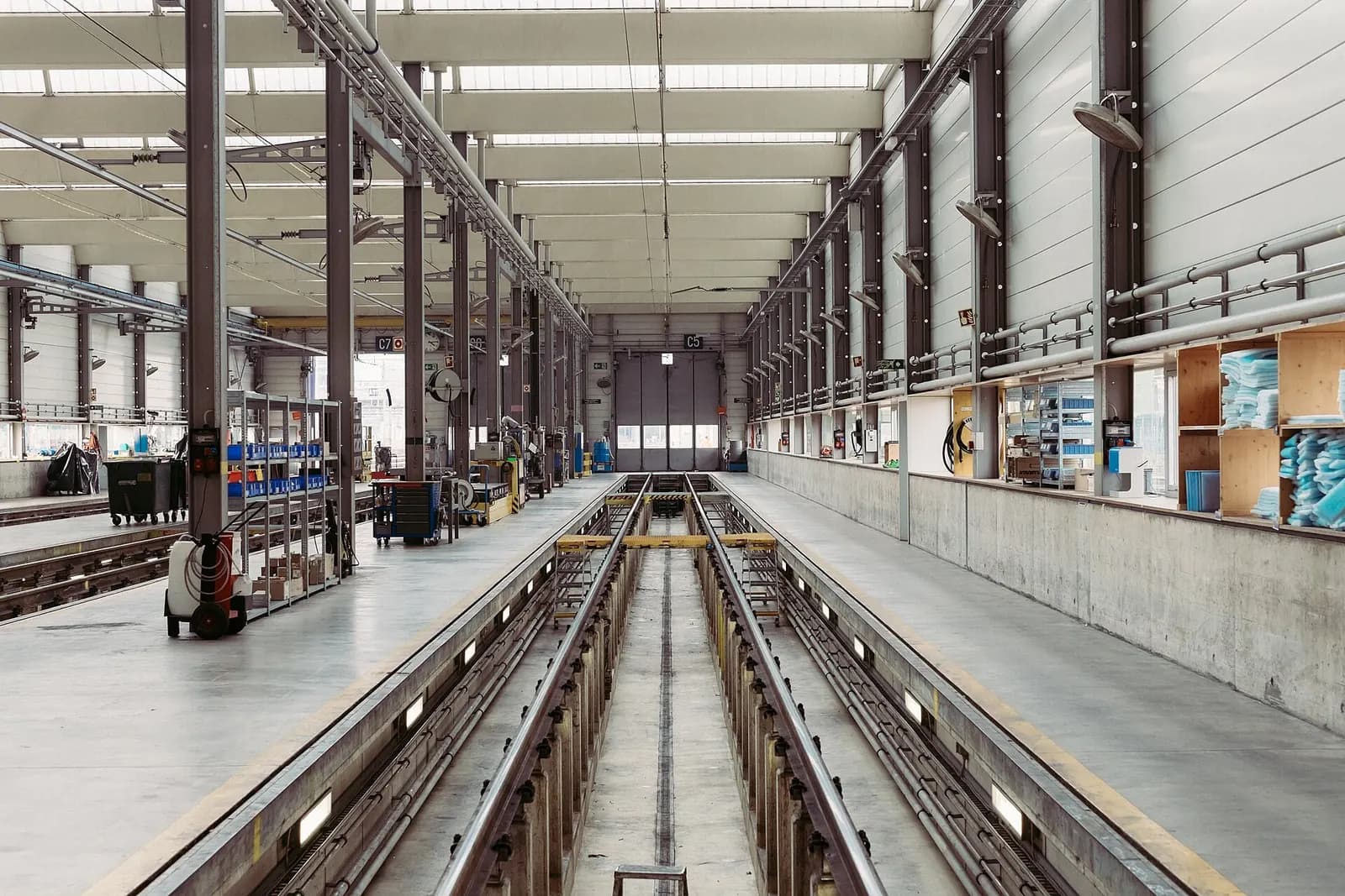 Long industrial maintenance hall with inspection pits between rails, overhead service lines and workbenches along the walls