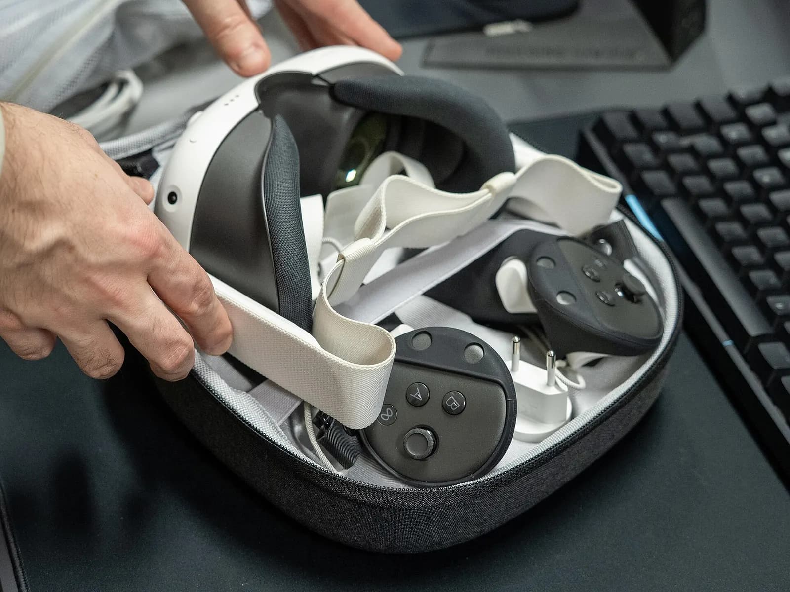 Hands holding a white standalone VR headset and its controllers beside a keyboard