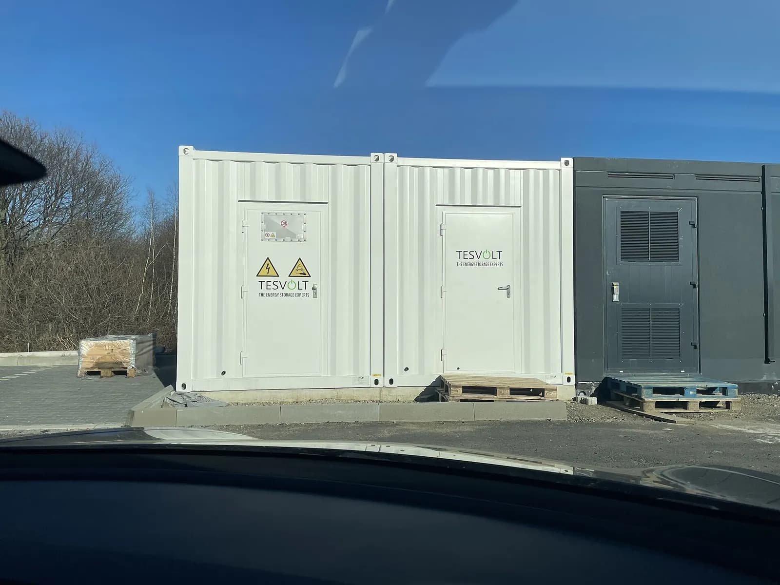 White containerised battery storage units with warning labels on a gravel site, seen through a vehicle windscreen