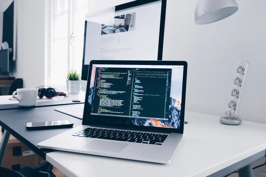 Laptop showing source code in a dark editor on a desk, with a monitor, phone, headphones and a coffee cup