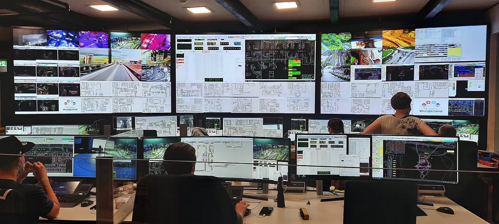 Control room with a wall of large displays showing track diagrams and camera feeds, and operators at desks with several monitors each