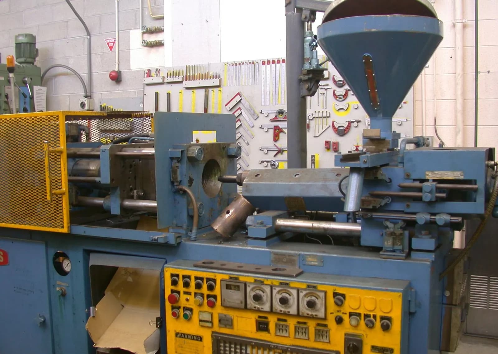 Older blue plastics injection moulding machine with a yellow control panel and a material hopper in a workshop