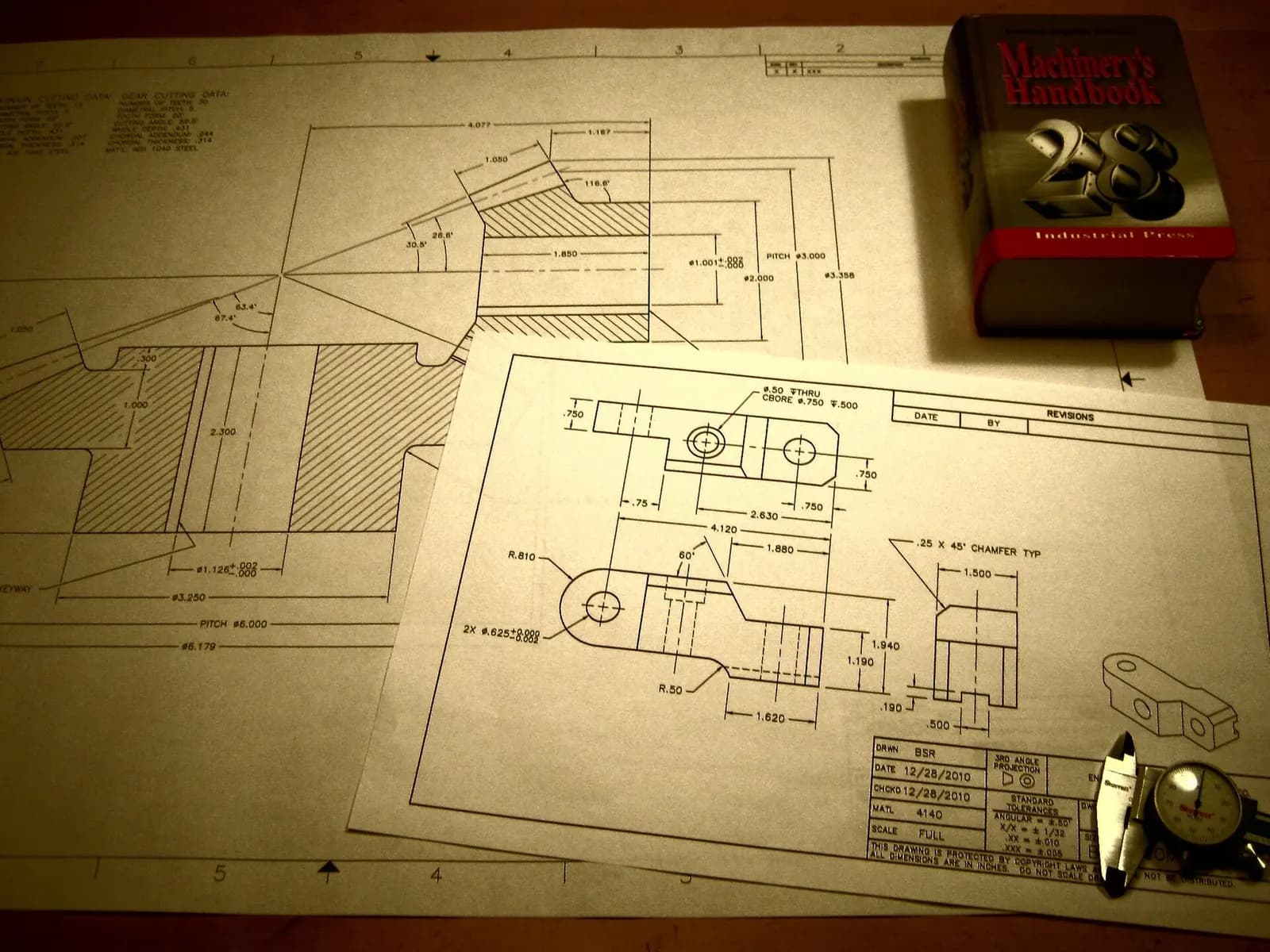 Printed engineering drawings with dimensions and a title block spread on a desk next to a Machinery's Handbook