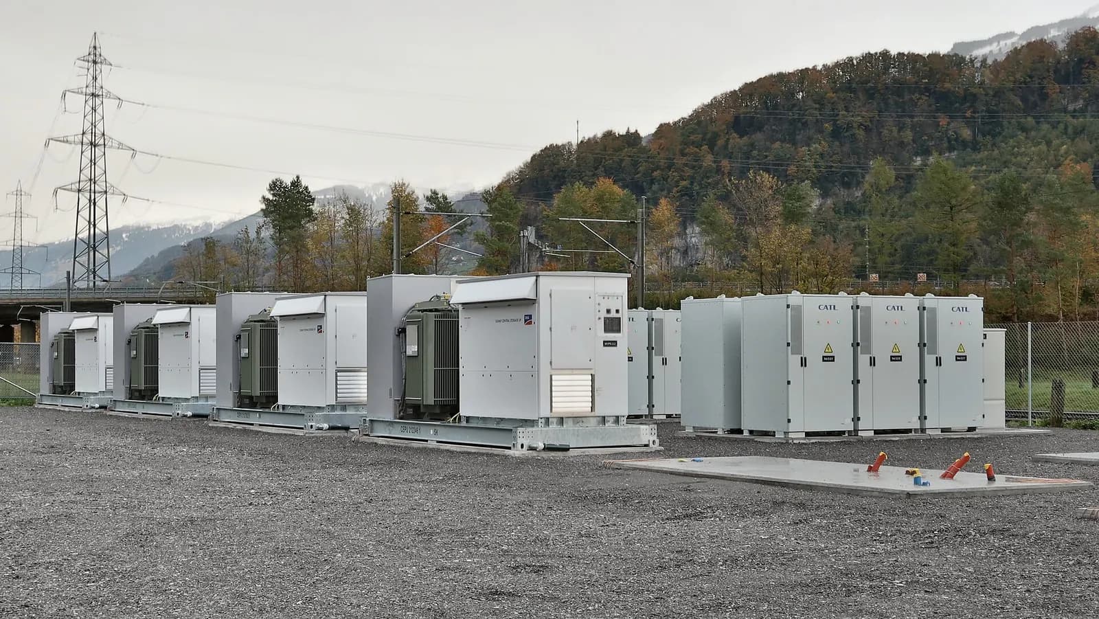 Row of white battery storage containers and transformer units at a grid battery site in a mountain valley