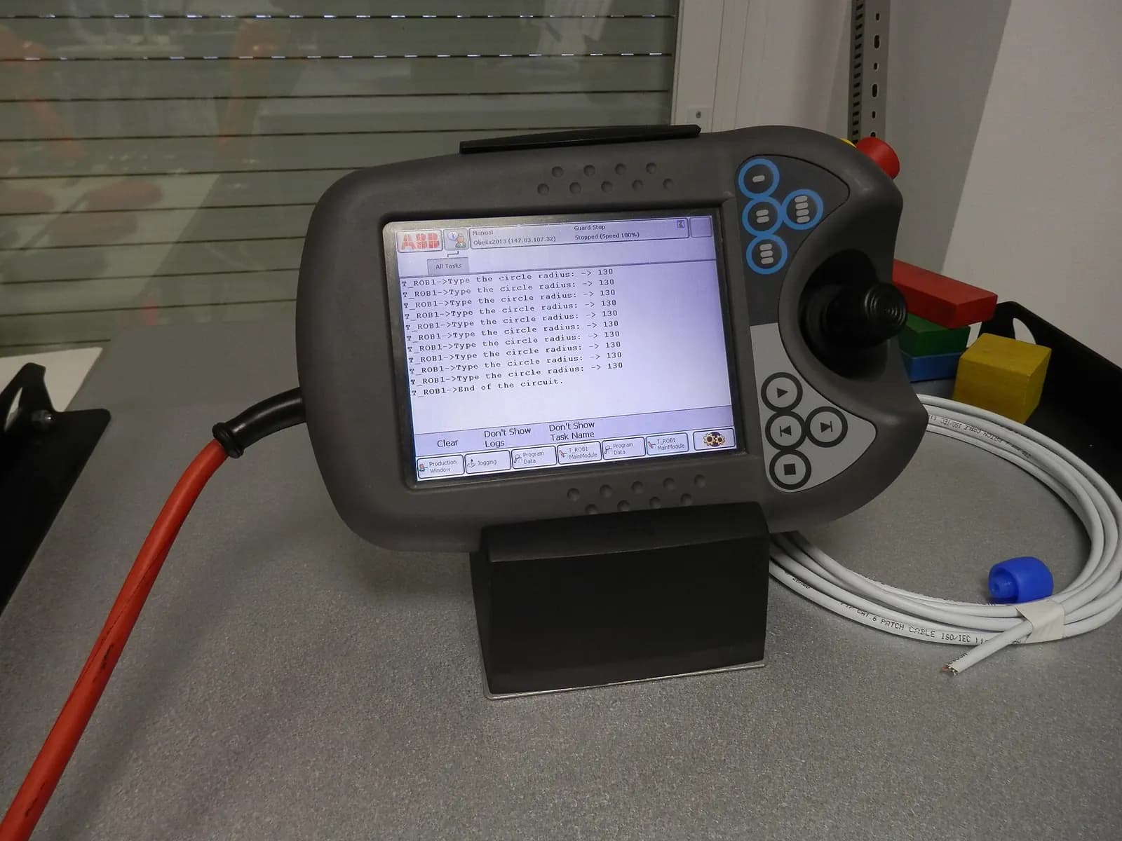 ABB robot teach pendant resting on a desk in a training room, connected by its cable