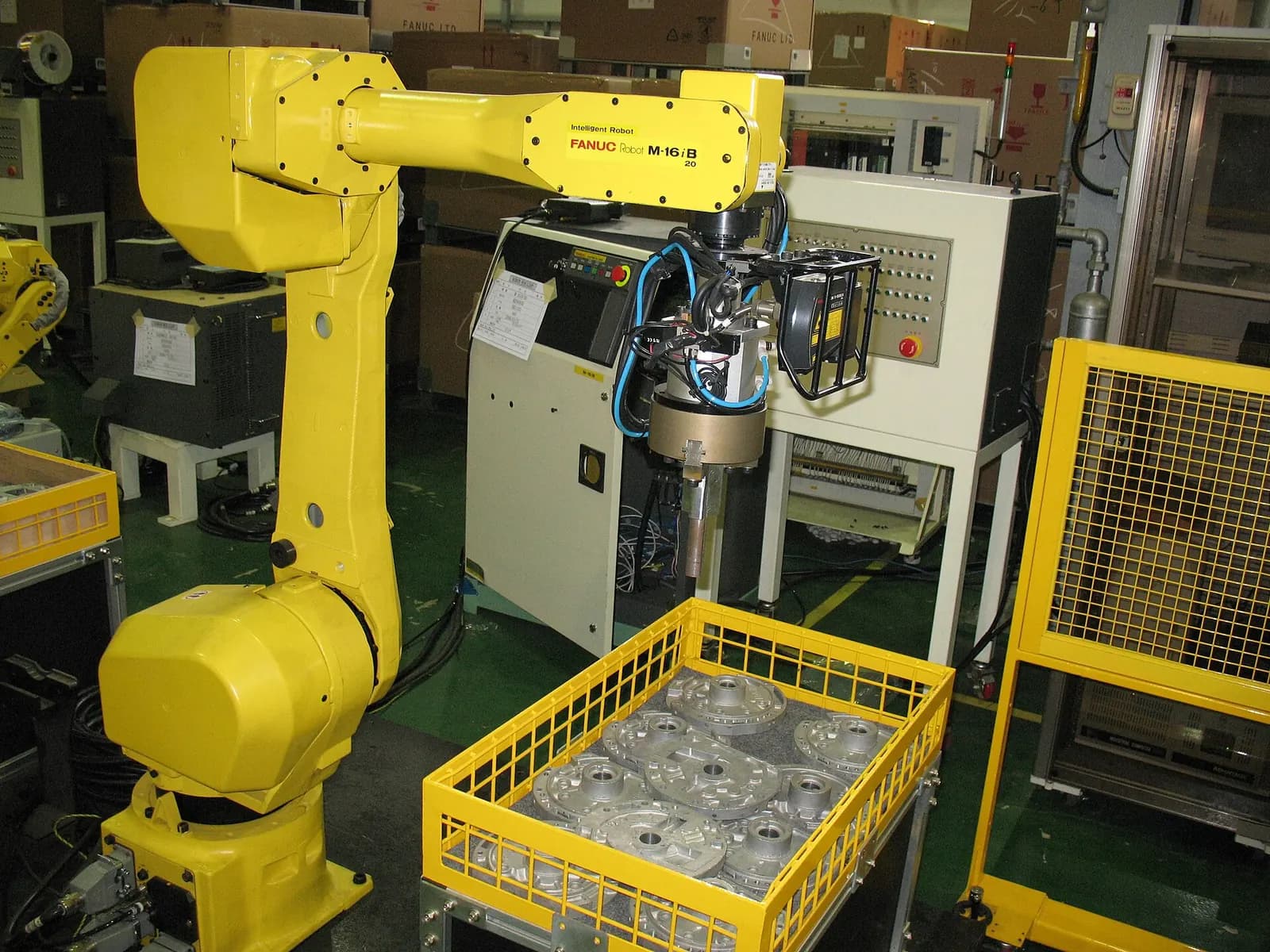Yellow FANUC industrial robot with a gripper beside a machine and a yellow crate of parts inside a guarded cell