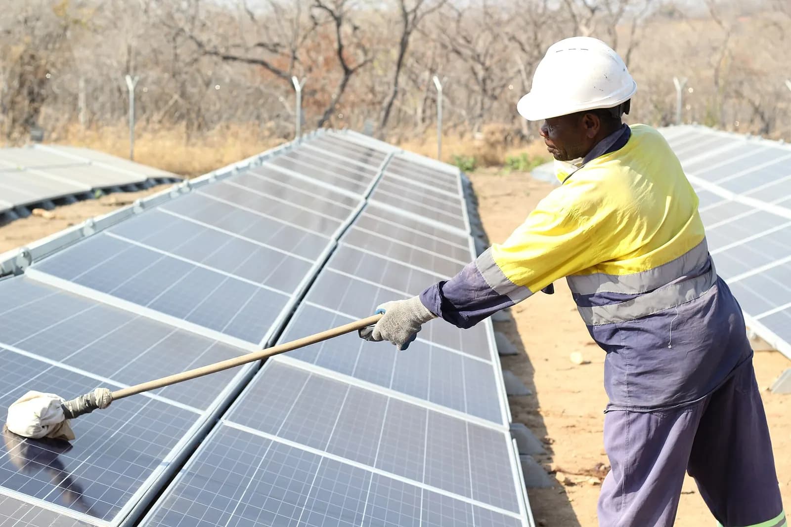 Worker in a hard hat cleaning a row of solar panels with a long-handled soft brush