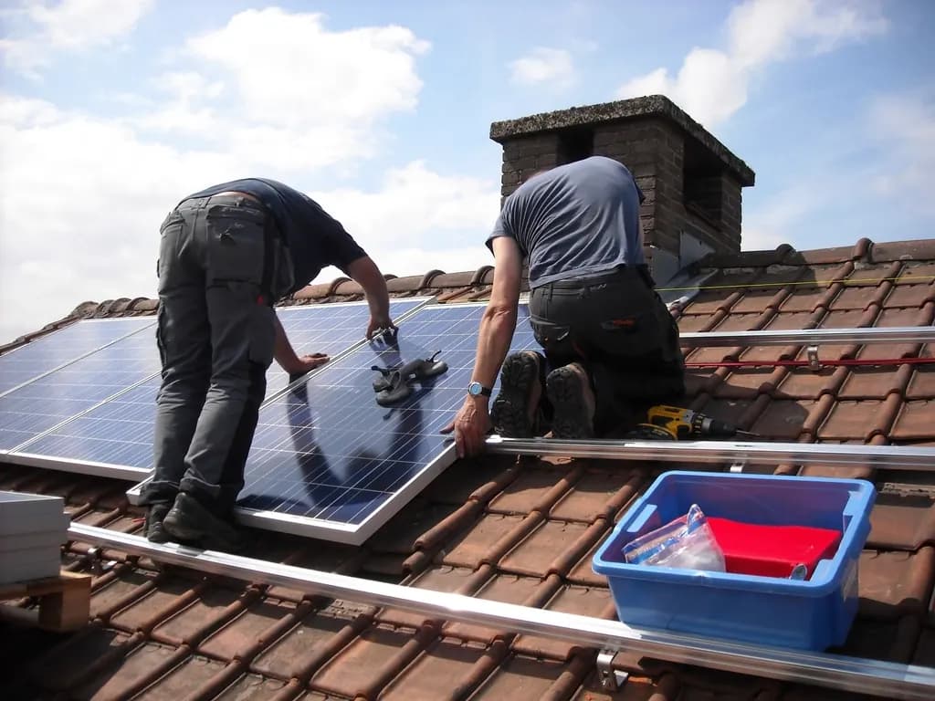 Two installers fixing solar panels onto mounting rails on a sloping clay-tiled roof under a blue sky
