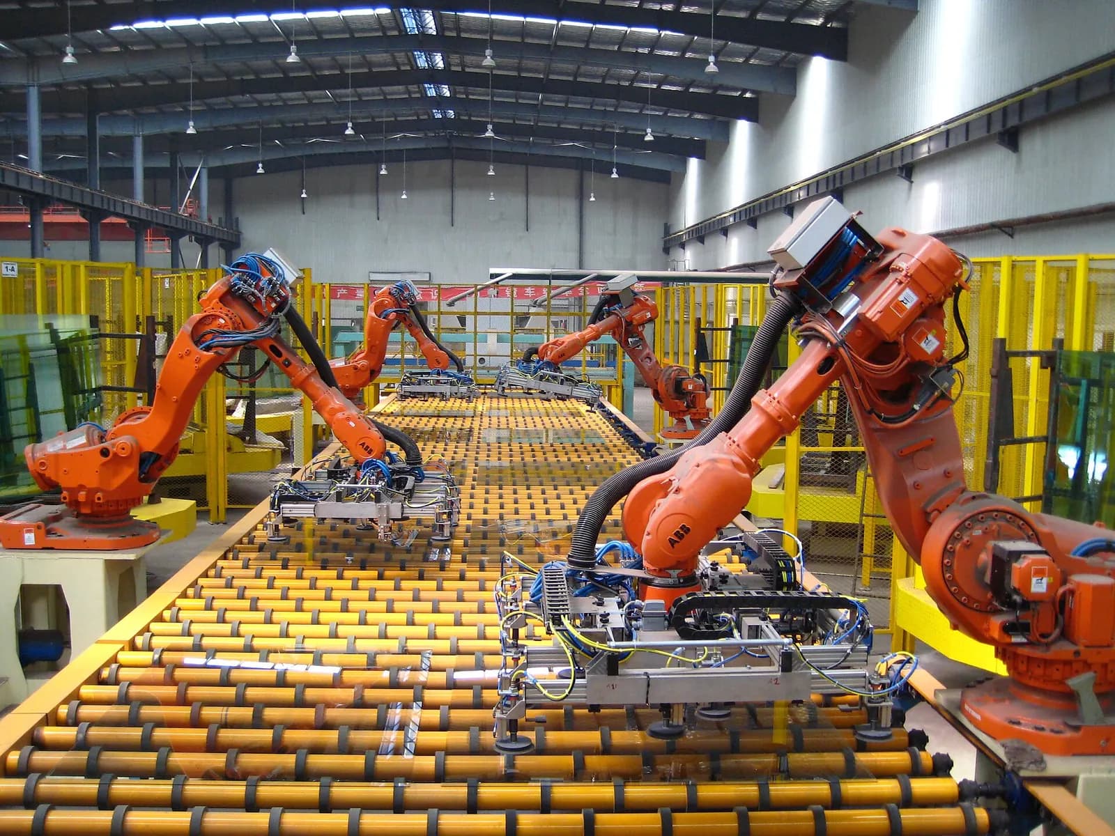 Several orange industrial robots with suction grippers unloading glass sheets from a long roller conveyor in a factory