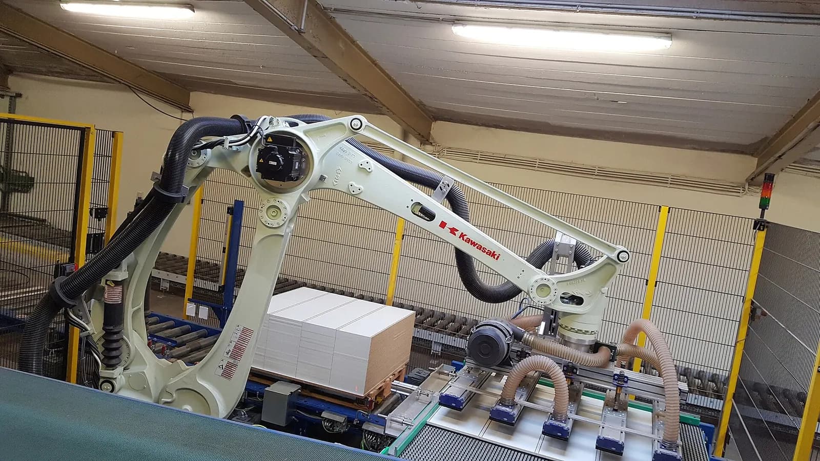 Robotic palletising cell with a large white robot arm and vacuum gripper stacking boards beside roller conveyors behind safety fencing