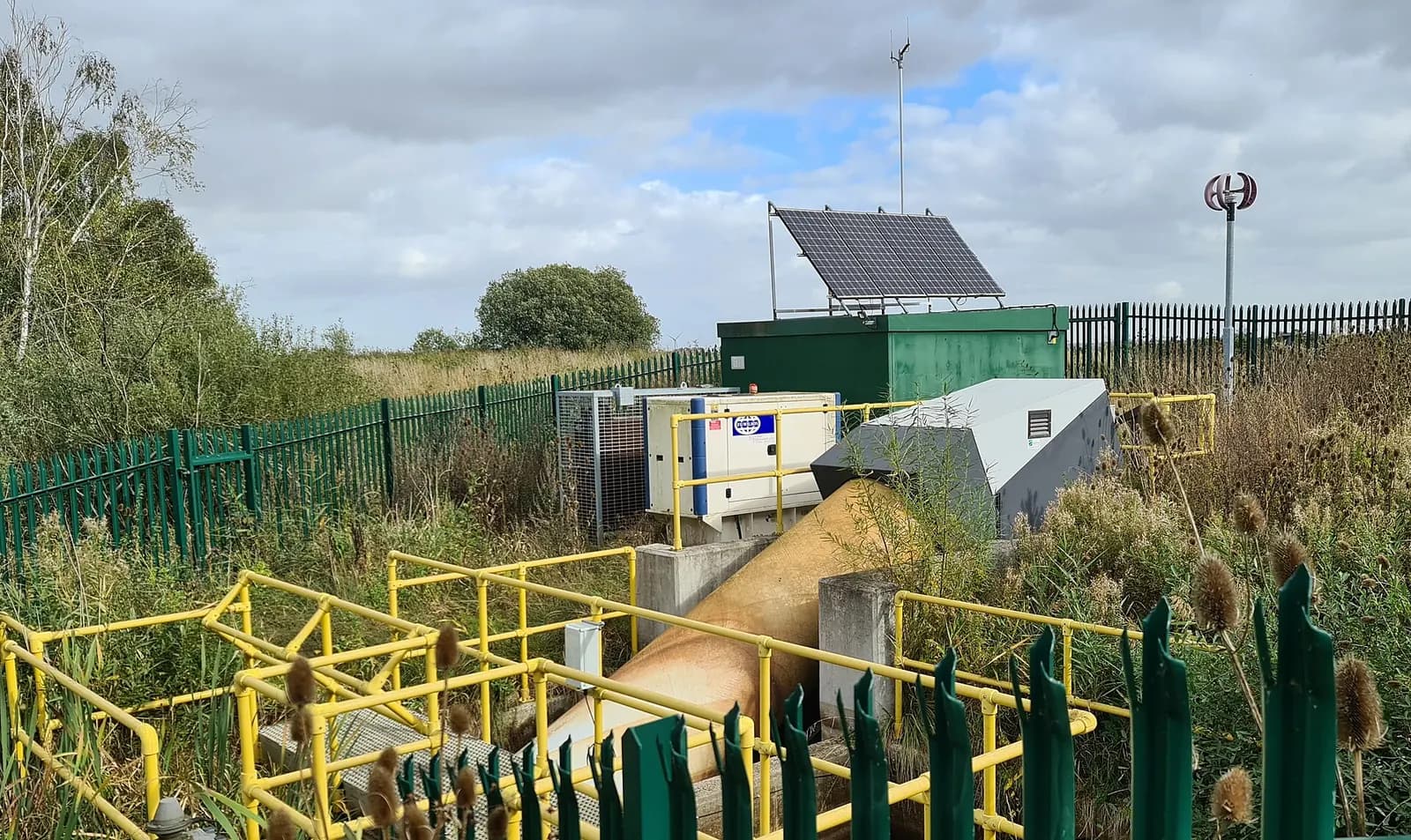 Small water pumping station compound with a green kiosk, solar panel and yellow handrails