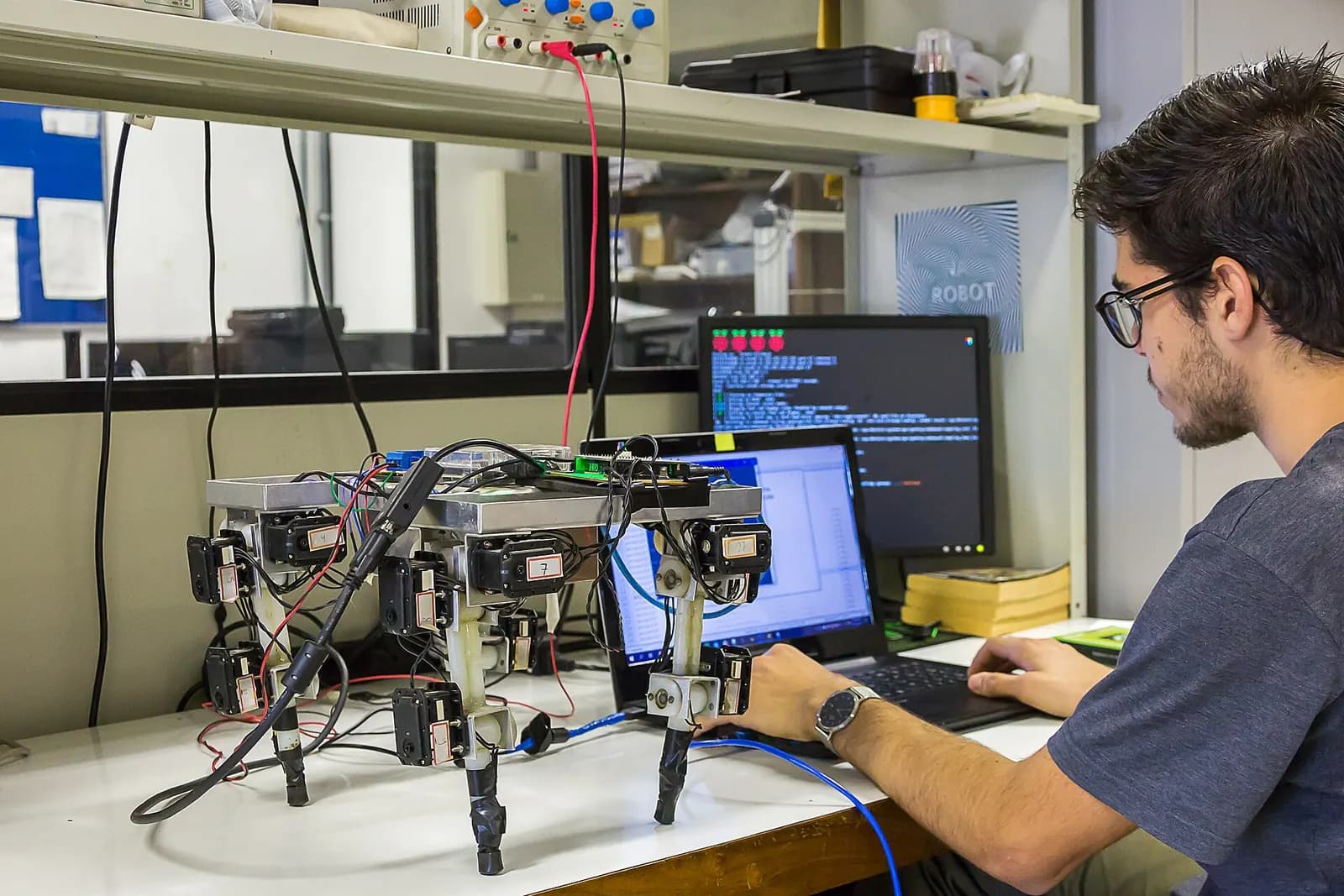 Engineer programming a small robotic rig from a laptop in a university laboratory