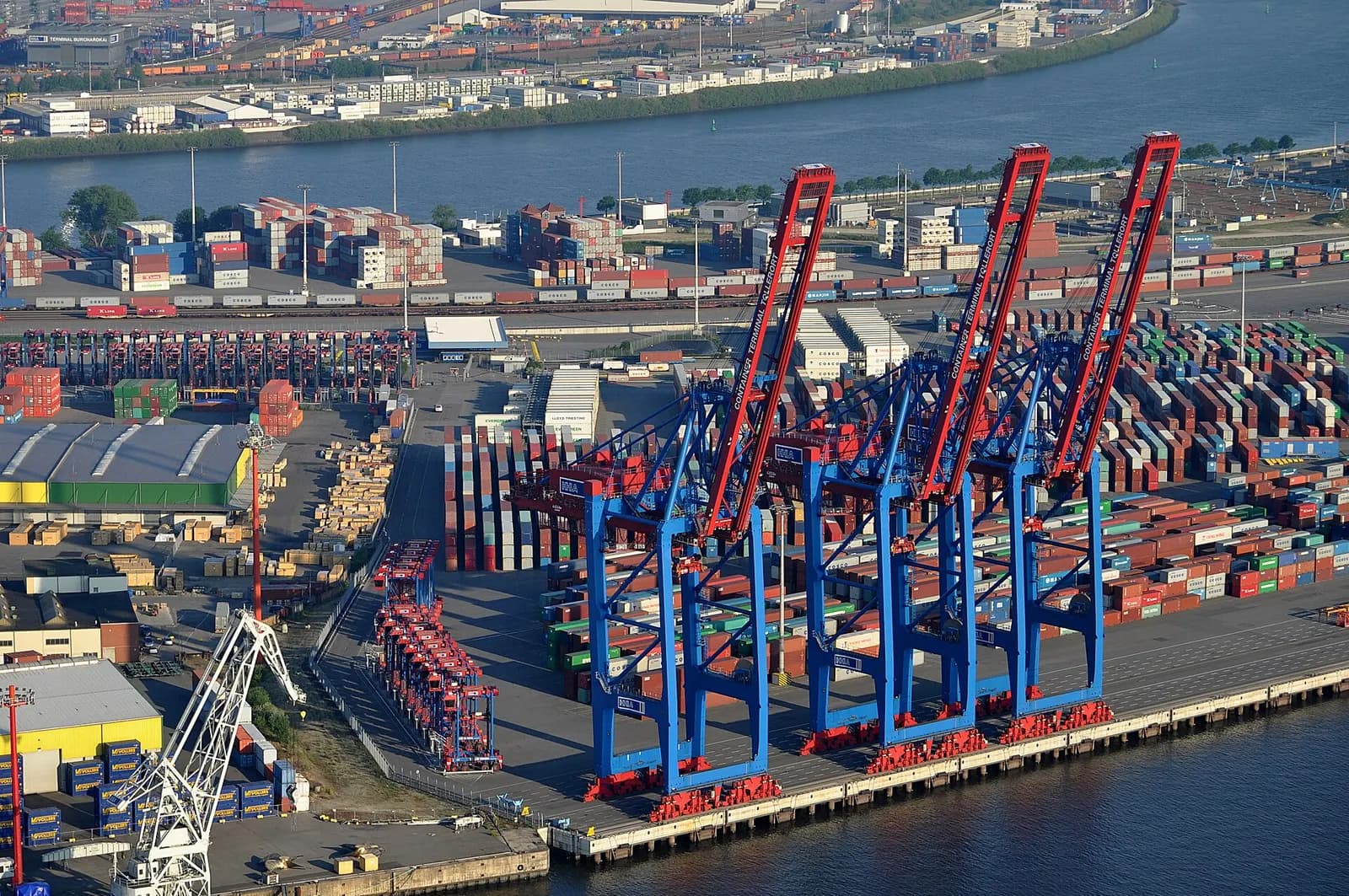 Aerial view of ship-to-shore container cranes and stacked containers at a container terminal on a river.