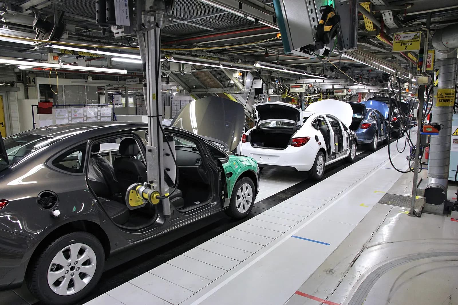 Car bodies with open doors moving along a final assembly line under overhead tooling in a car factory.
