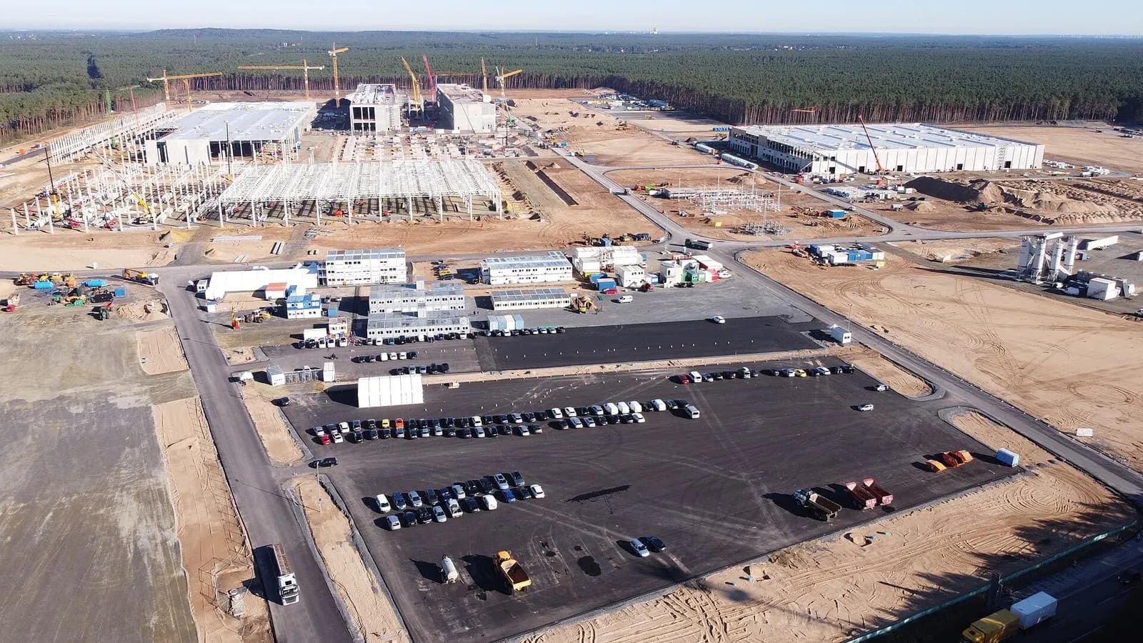 Aerial view of a large factory under construction with steel frames, cranes, site cabins and a car park.