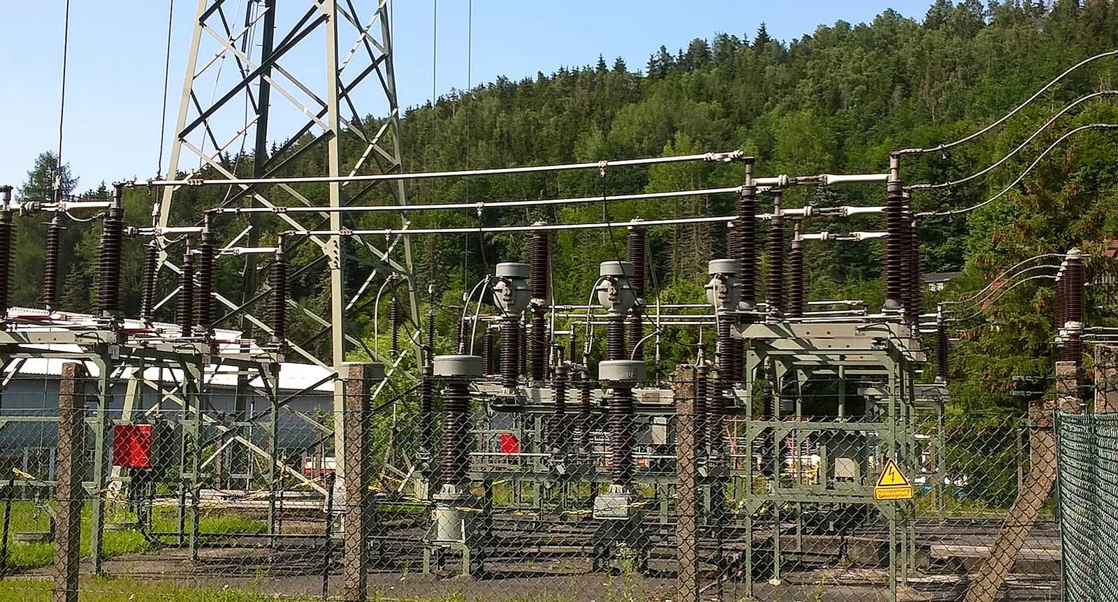 High-voltage substation with circuit breakers, disconnectors, transformers and gantries below a transmission tower.