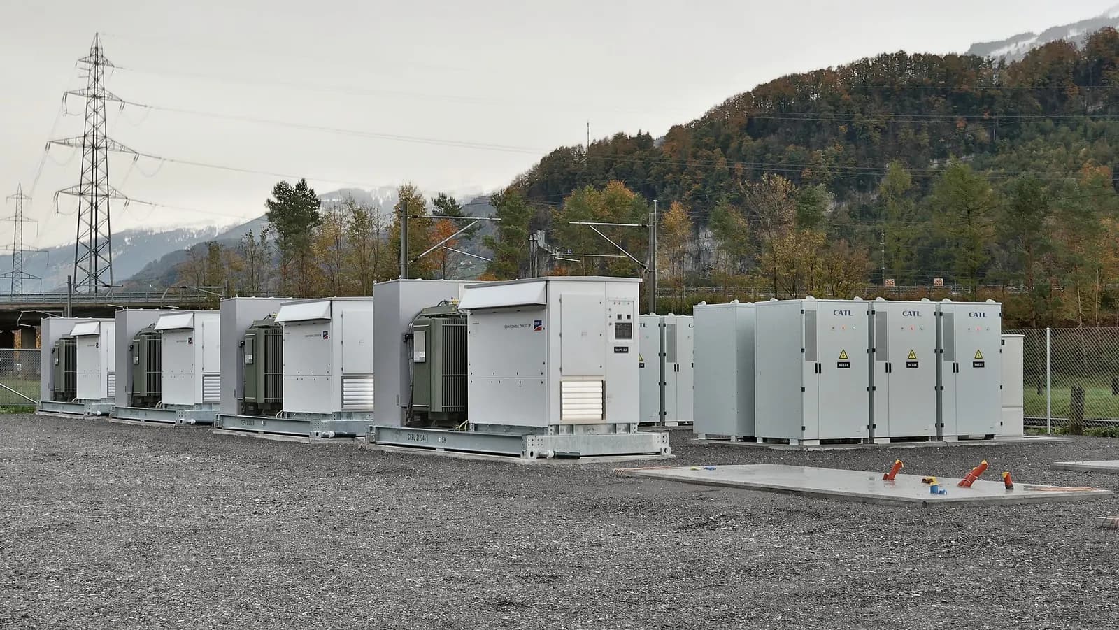 Containerised battery energy storage units, inverters and transformers on a gravel site beside a high-voltage line.
