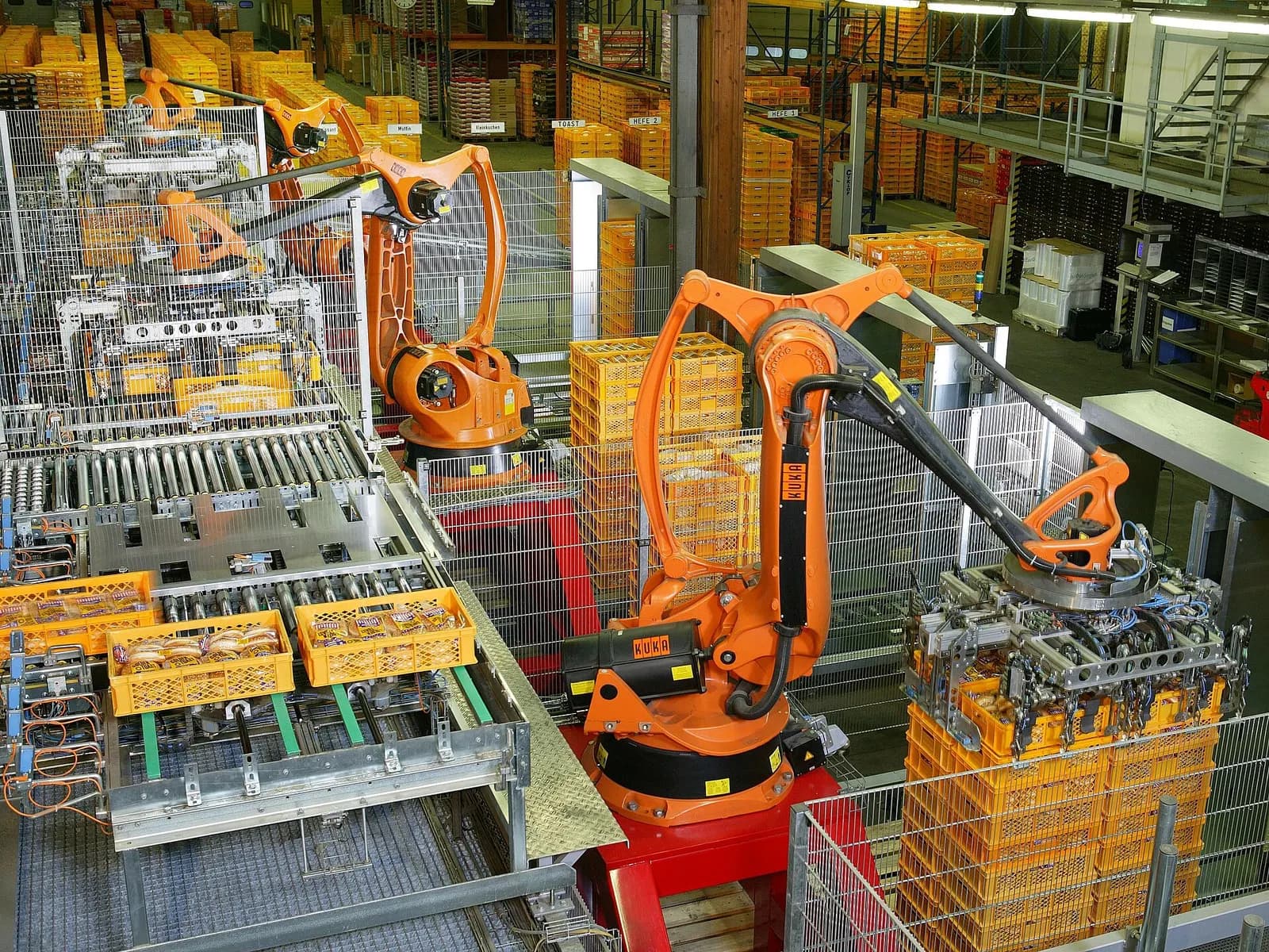 Orange industrial robots palletising crates of bread from roller conveyors at the end of a bakery packaging line.