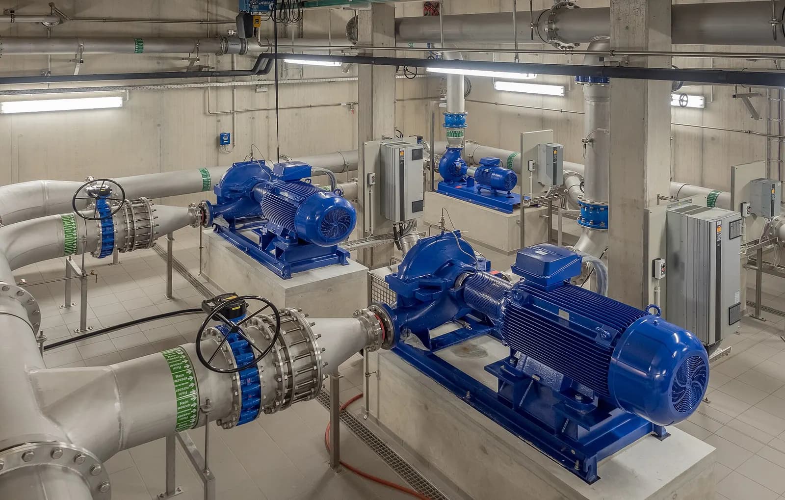 Pump hall in a waterworks with blue split-case pumps and motors connected to large stainless steel pipework and valves.