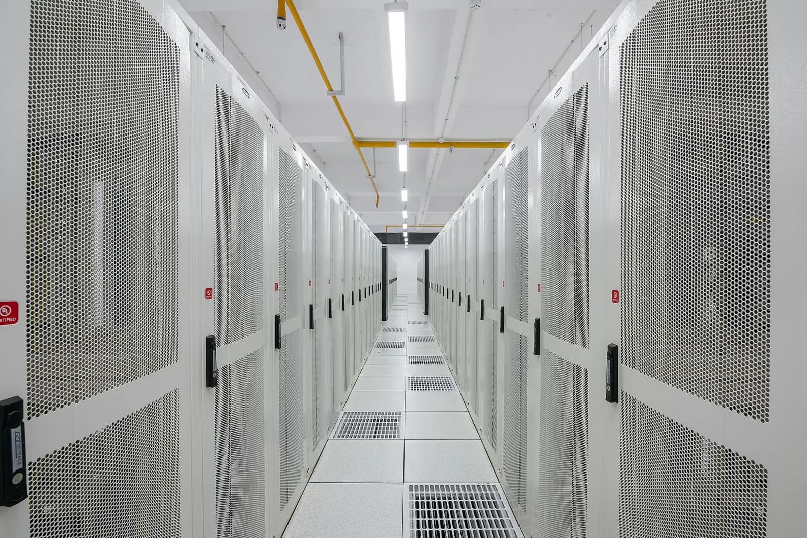 A cold aisle in a data hall lined with rows of white perforated server rack cabinets on a raised floor.